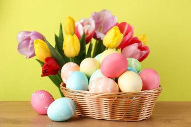 Hasır sepetin içine boyanmış Paskalya yumurtaları ahşap masanın üzerinde ve sarı arka planda güzel laleler, yakın plan.