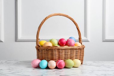 Hasır sepete boyanmış Paskalya yumurtaları beyaz duvarın yanındaki beyaz mermer masada.