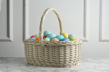 Hasır sepete boyanmış Paskalya yumurtaları beyaz duvarın yanındaki beyaz mermer masada.