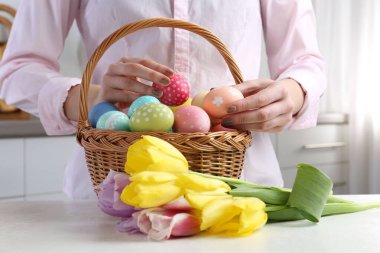 Paskalya sepetinde yumurta boyalı bir kadın ve beyaz masa içinde laleler, yakın plan.