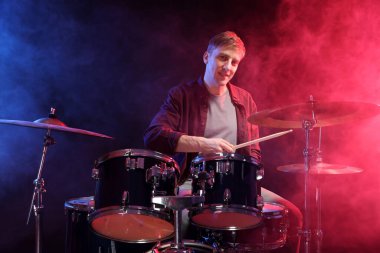 Gülümseyen adam davul çalıyor. Renkli ışıklar ve siyah arka planda duman.