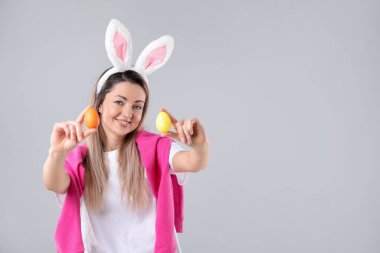 Paskalya kutlaması. Saçbantlı, tavşan kulaklı, gri arka planda boyanmış yumurta tutan gülümseyen bir kadın portresi.
