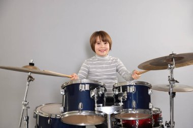 Açık gri arka planda bagetleri ve davulları olan küçük bir çocuk.