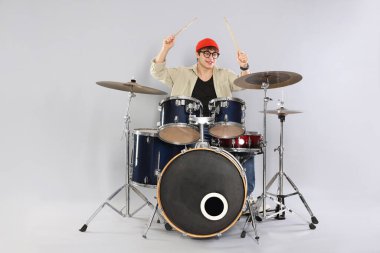 Gri arka planda davul çalmayı öğrenen, gözlüklü ve şapkalı gülümseyen genç çocuk.