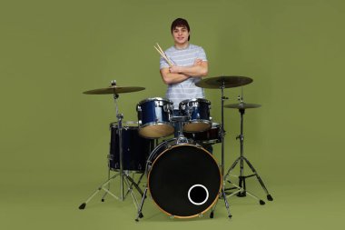 Beyzbol şapkalı, davul setiyle yeşil arka planda gülümseyen genç çocuk.