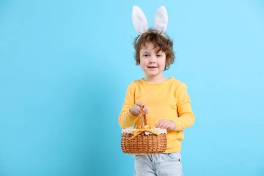 Paskalya kutlaması. Saçbantlı, tavşan kulaklı ve açık mavi arka planda boyalı yumurta dolu sepeti olan gülümseyen küçük bir çocuk. Metin için boşluk