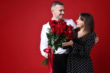 Sevgililer Günü kutlaması. Erkek, kırmızı arka planda gül buketiyle sevgilisine sürpriz yapıyor. Mesaj için yer var.