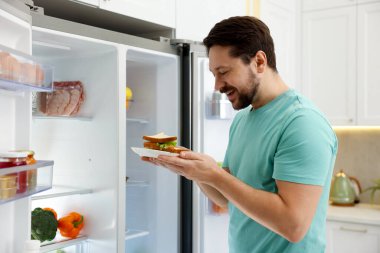 Mutfaktaki açık buzdolabının yanında sandviçleri olan adam.