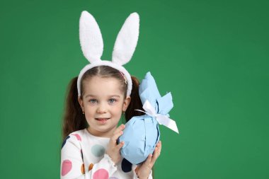 Paskalya kutlaması. Saçbantlı, tavşan kulaklı gülümseyen küçük bir kızın portresi yeşil arka planda sarılı bir hediye, mesaj için yer var.