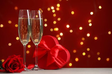 Sevgililer Günü. Köpüklü şarap kadehleri, hediye kutusu ve ahşap masanın üzerinde kırmızı arka planda bulanık ışıklar ve yazı için yer olan gül. Bokeh etkisi