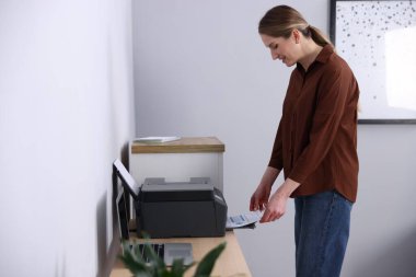 Gri duvarın yanındaki yazıcıdan baskı kağıdı alan gülümseyen kadın.