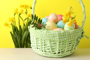 Hasır sepetin içine boyanmış Paskalya yumurtaları bej masa üzerinde ve nergisler sarı arka planda, yakın plan