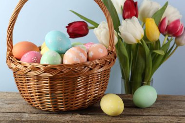 Hasır sepetin içine boyanmış Paskalya yumurtaları ahşap masada ve açık mavi arka planda güzel laleler, yakın planda.