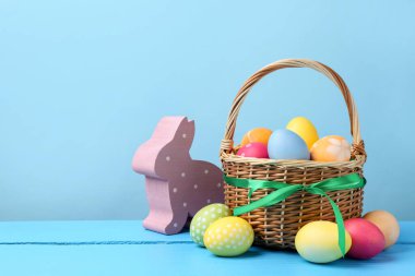 Hasır sepete boyanmış Paskalya yumurtaları ve açık mavi arka planda ahşap masa üzerindeki dekoratif tavşan figürü. Metin için boşluk
