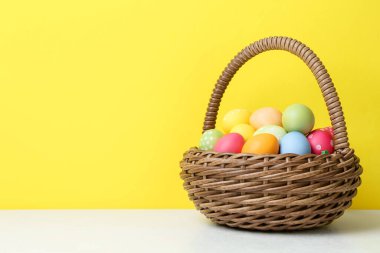 Hasır sepetin içine boyanmış Paskalya yumurtaları beyaz masada, sarı arka planda. Metin için boşluk