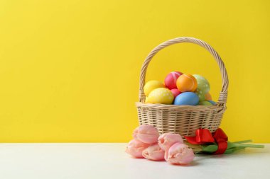 Hasır sepete boyanmış Paskalya yumurtaları ve sarı arka planda beyaz masada güzel laleler. Metin için boşluk