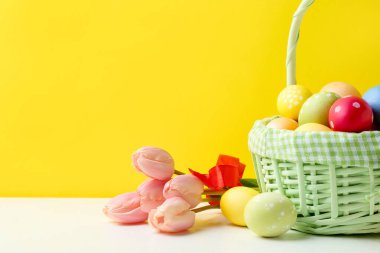 Hasır sepete boyanmış Paskalya yumurtaları ve sarı arka planda beyaz masada güzel laleler. Yakın plan. Metin için boşluk