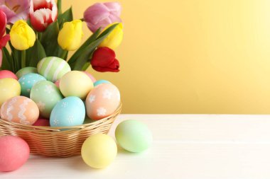 Hasır sepete boyanmış Paskalya yumurtaları beyaz ahşap masa üzerinde ve güzel laleler sarı arka planda, yakın plan. Metin için boşluk