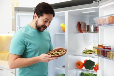 Mutfakta açık buzdolabının yanında turtası olan bir adam.