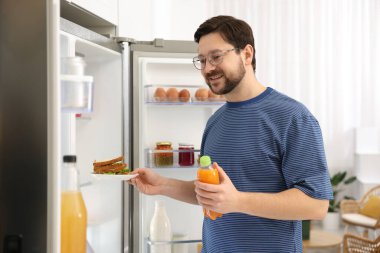 Mutfakta açık buzdolabının yanında sandviç ve meyve suyu olan bir adam.