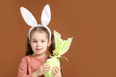 Paskalya kutlaması. Saçbandı takmış, tavşan kulaklı gülümseyen küçük bir kızın portresi kahverengi arka planda sarılı bir hediye, mesaj için yer var.