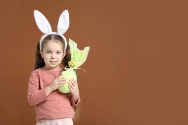 Paskalya kutlaması. Saçbandı takmış, tavşan kulaklı gülümseyen küçük bir kızın portresi kahverengi arka planda sarılı bir hediye, mesaj için yer var.