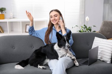 Akıllı telefonuyla gülümseyen bir kadın ve evdeki kanepede oturan sevimli köpeği.