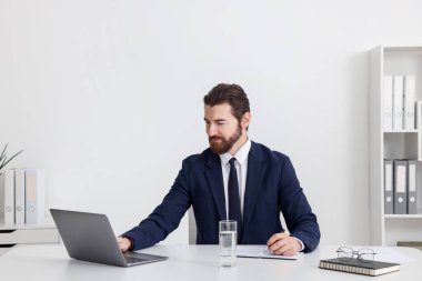 Beyaz duvarın yanındaki masada dizüstü bilgisayarla çalışan bir iş adamı.