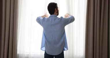 Adam evde, arka planda şık pencere perdeleri açıyor