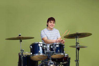 Beyzbol şapkalı gülümseyen genç, bagetleriyle yeşil arka planda davul çalmayı öğreniyor.