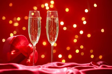 Sevgililer Günü. Şarap kadehleri ve hediye kutusu kırmızı arka planda ipeğe karşı bulanık ışıklarla, yakın plan. Metin için boşluk