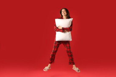 Kırmızı arka planda yastıkla zıplayan şık pijamalı mutlu kadın.