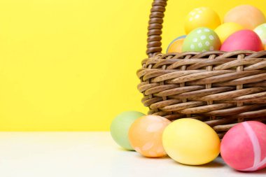 Hasır sepetin içine boyanmış Paskalya yumurtaları beyaz masada, sarı arka planda, yakın plan. Metin için boşluk