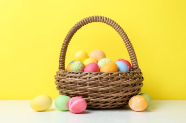 Hasır sepetin içine boyanmış Paskalya yumurtaları beyaz masada sarı arka planda.