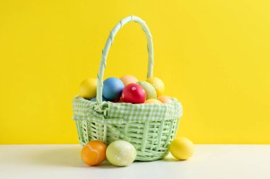 Hasır sepetin içine boyanmış Paskalya yumurtaları beyaz masada sarı arka planda.