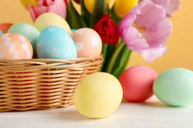 Hasır sepetin içine boyanmış Paskalya yumurtaları beyaz ahşap masa üzerinde ve sarı arka planda güzel laleler, yakın plan.