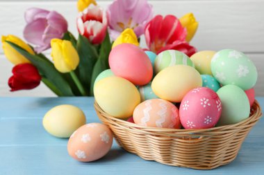 Hasır sepetin içine boyanmış Paskalya yumurtaları açık mavi ahşap masa ve kapalı alanda güzel laleler. Yakın plan.