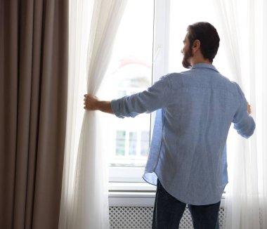 Adam evde, arka planda şık pencere perdeleri açıyor