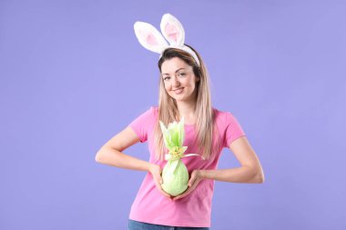 Paskalya kutlaması. Saç bandı takmış, tavşan kulaklı, gülümseyen bir kadın menekşe arka planda hediyesini tutuyor.