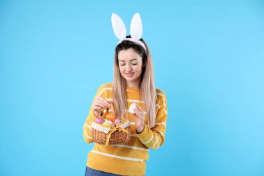 Paskalya kutlaması. Saçbantlı, tavşan kulaklı ve açık mavi arka planda boyalı yumurtalarla dolu sepeti olan gülümseyen kadın.