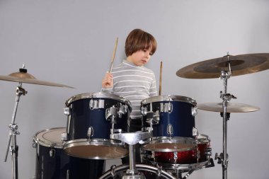 Açık gri arka planda bagetleri ve davulları olan küçük bir çocuk.