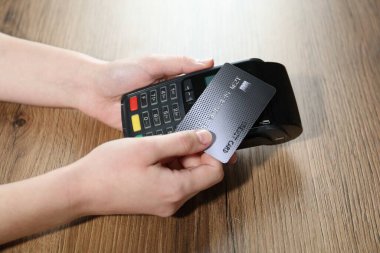 Kredi kartı olan bir kadın ahşap masada ödeme terminalini kullanıyor, yakın plan.