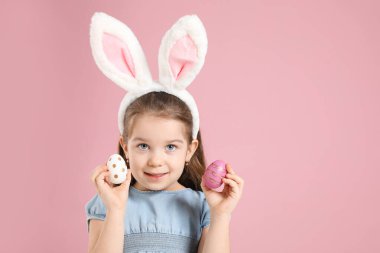 Paskalya kutlaması. Saçbantlı, tavşan kulaklı, pembe arka planda boyalı yumurtalar tutan gülümseyen küçük bir kızın portresi.