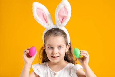 Paskalya kutlaması. Sarı arka planda boyalı yumurtalar tutan, saç bandı takmış, gülümseyen küçük bir kızın portresi.