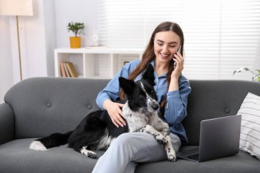 Akıllı telefonuyla gülümseyen bir kadın ve evdeki kanepede oturan sevimli köpeği.