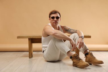 Moda modeli. Şapkalı ve güneş gözlüklü yakışıklı adam bej arka planda bankın yanında poz veriyor.