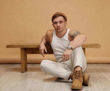 Moda modeli. Şapkalı yakışıklı adam bej arka plana yaslanarak yerde poz veriyor.