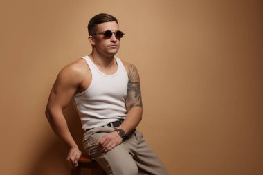 Moda modeli. Güneş gözlüklü yakışıklı bej arka planda taburede poz veriyor, mesaj için yer var.