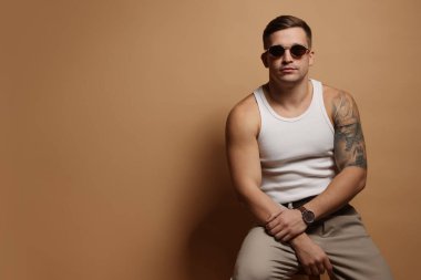 Moda modeli. Güneş gözlüklü yakışıklı bej arka planda taburede poz veriyor, mesaj için yer var.