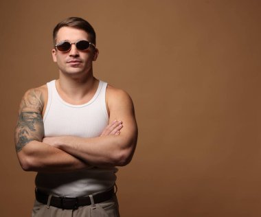 Moda modeli. Güneş gözlüklü yakışıklı bej arka planda çapraz kollarla poz veriyor, mesaj için yer var.
